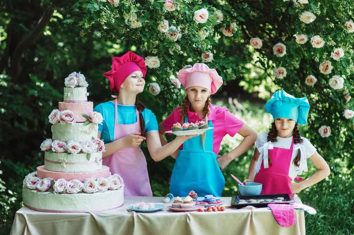 Three girls in chef hats and aprons at a table with a tiered cake and cupcakes