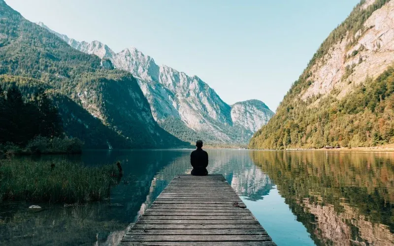 A person sitting alone at the end of a dock, facing a mountain lake