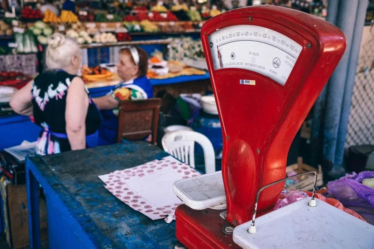 A red mechanical scale on a blue market counter, two women at a stall behind