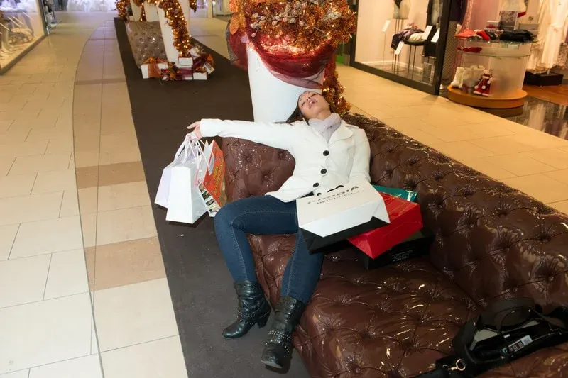 An exhausted woman slumps on a sofa in a mall, surrounded by shopping bags