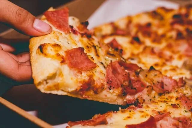 Kawałek pizzy z wędliną i serem trzymany w dłoni nad pudełkiem