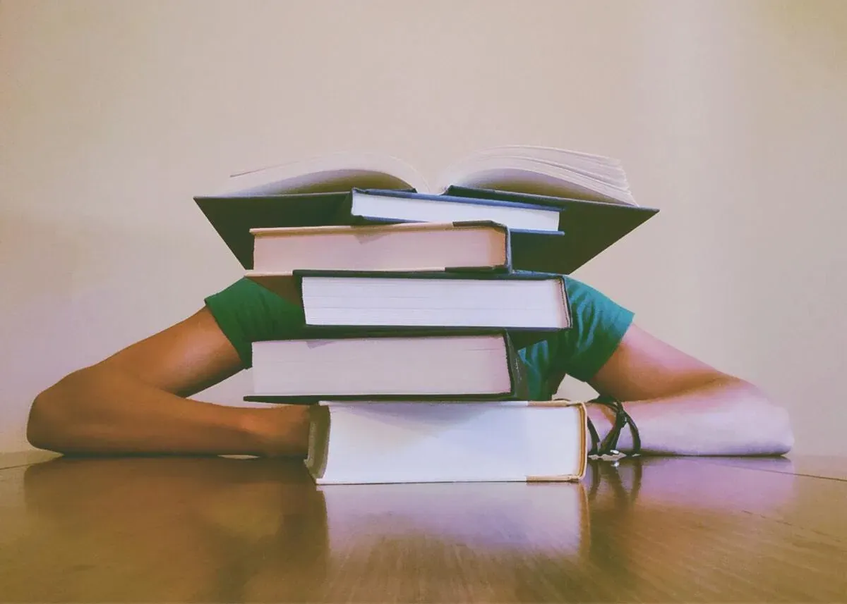 A student hides behind a tall stack of hardcover books, an open book on top