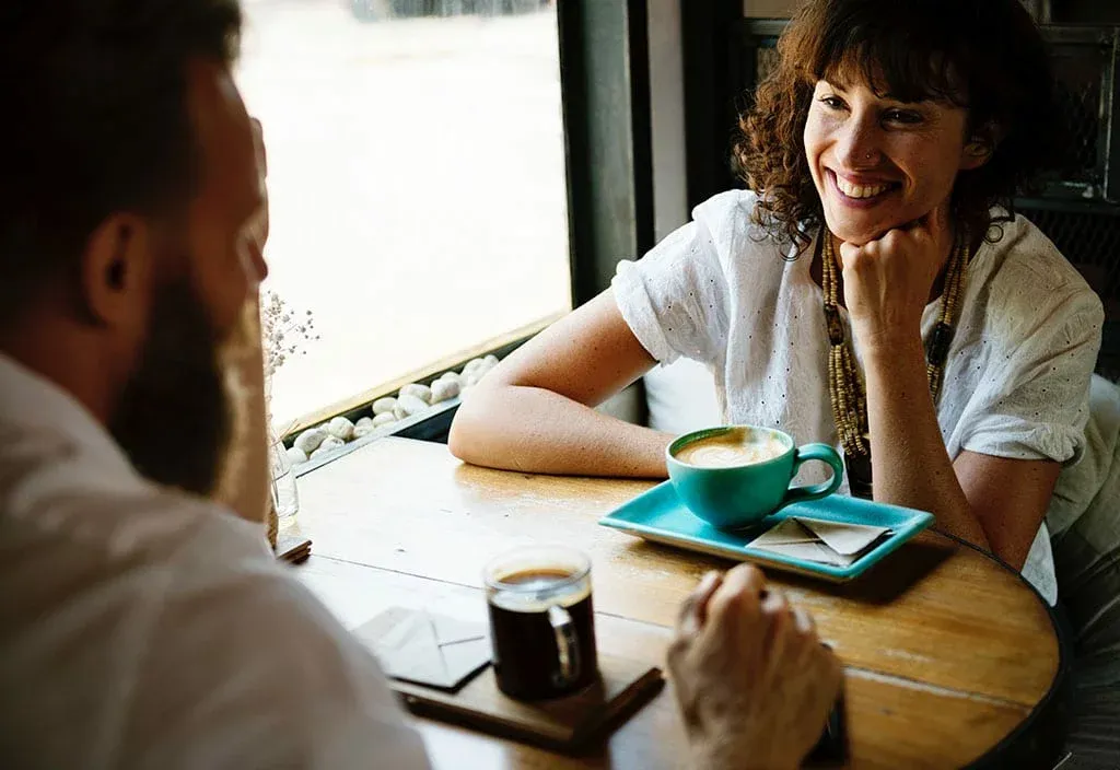 木のカフェテーブルで、笑顔の女性がひげの男性とコーヒーを飲みながら話している